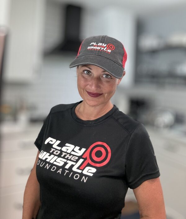 A woman wearing a "Play to the Whistle Foundation" hat and matching T-shirt stands in a modern kitchen.