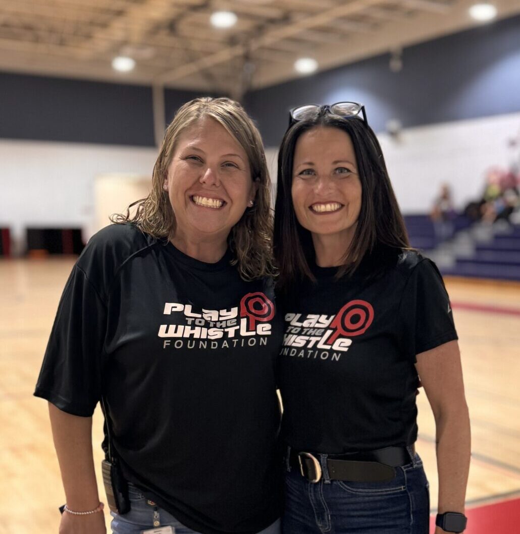 Two women stand smiling in a gymnasium, wearing matching "Play Whistle Foundation" Ladies T-Shirts. One holds a microphone and the other has keys in her hand.