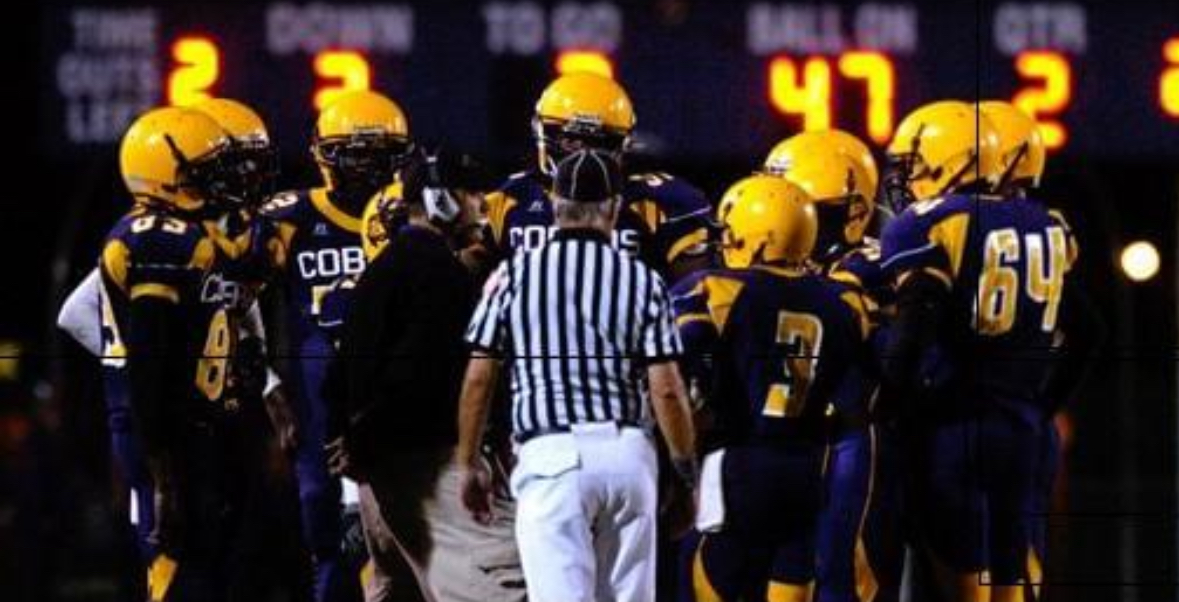 A group of American football players in yellow helmets and blue uniforms gather around a referee on the field during a game. The scoreboard in the background displays "Down 2," "To Go 1," and "Ball on 47.